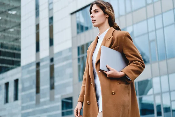 Молодая серьезная деловая женщина в пальто с ноутбуком задумчиво прогуливаясь по городской улице — стоковое фото