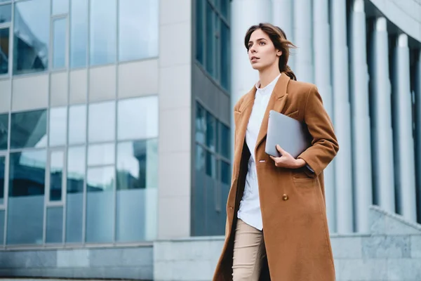 Young attractive businesswoman in coat with laptop confidently walking through city street — ストック写真
