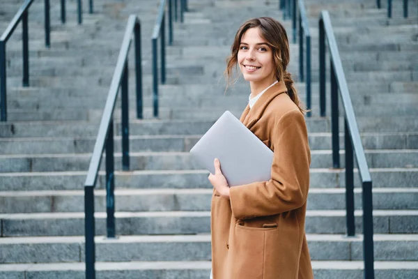 Giovane donna d'affari piuttosto casual in cappotto con computer portatile felicemente alla ricerca in macchina fotografica all'aperto — Foto Stock