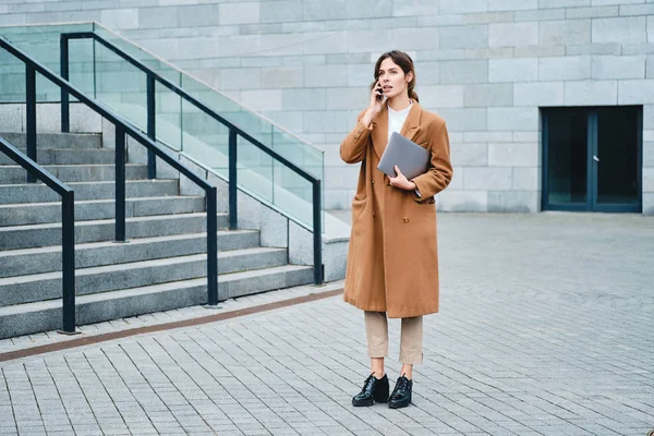 Jeune femme d'affaires attrayante en manteau avec ordinateur portable parler en toute confiance sur téléphone portable sur la rue de la ville — Photo