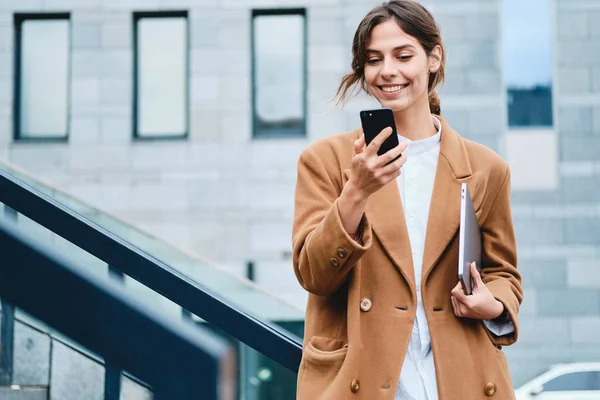 Wanita bisnis muda yang santai bermantel dengan laptop dengan senang hati menggunakan ponsel di luar — Stok Foto