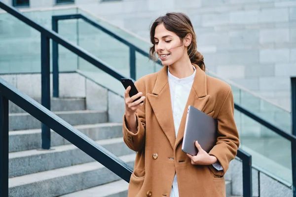 Молода усміхнена випадкова бізнес-леді в пальто з ноутбуком щасливо використовує мобільний телефон на відкритому повітрі — стокове фото
