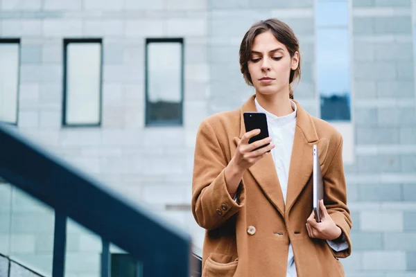 Молодая серьезная деловая женщина в пальто с ноутбуком уверенно используя мобильный телефон на открытом воздухе — стоковое фото