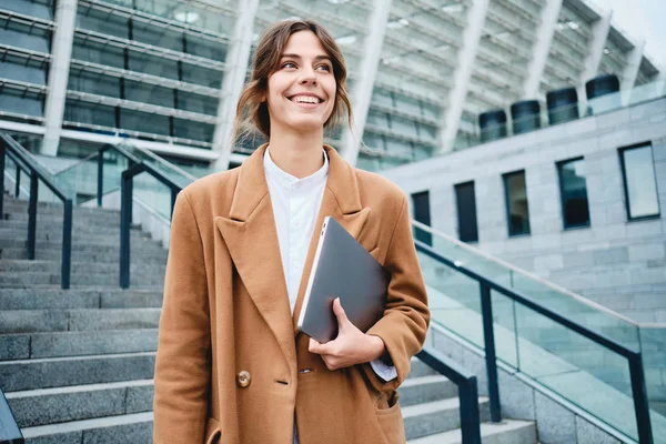 Giovane donna d'affari piuttosto positivo in cappotto con il computer portatile gioiosamente guardando lontano all'aperto — Foto Stock