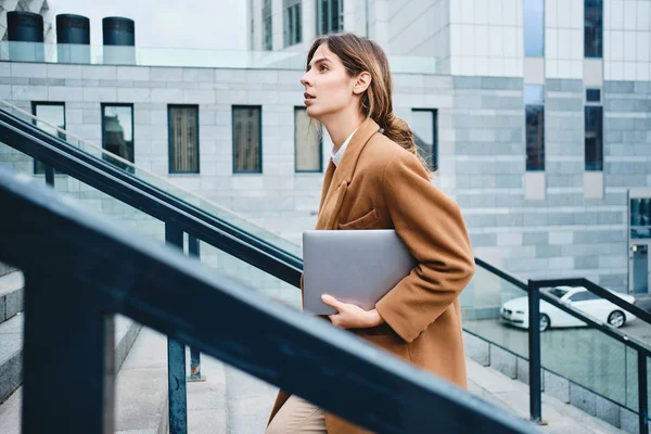 Вид сбоку молодой красивой бизнесвумен в пальто с ноутбуком, поднимающейся по лестнице на улицу — стоковое фото