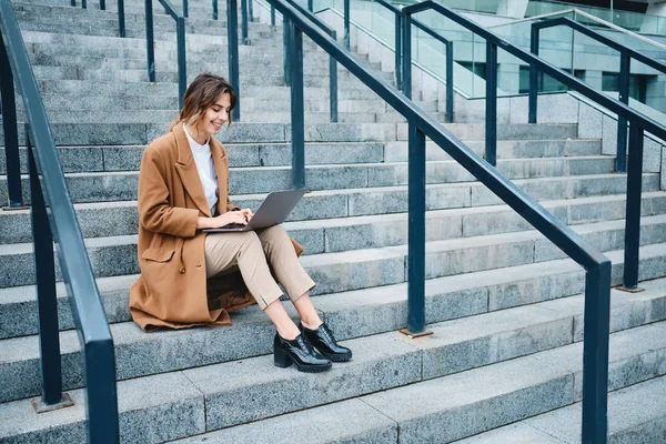 Jovem mulher de negócios muito sorridente no casaco feliz trabalhando no laptop em escadas ao ar livre — Fotografia de Stock