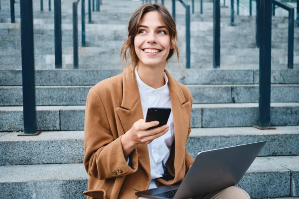 Jovem mulher de negócios bonita no casaco com celular e laptop alegremente olhando para longe nas escadas ao ar livre — Fotografia de Stock