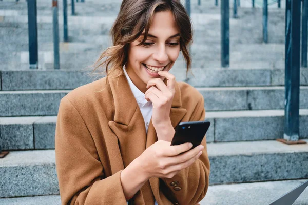 Portrett av en ung, opphisset forretningskvinne i frakk som med glede bruker mobiltelefon utendørs – stockfoto