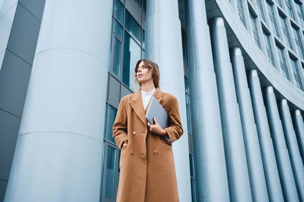 Jovem mulher de negócios elegante confiante no casaco com laptop olhando atentamente para o exterior — Fotografia de Stock