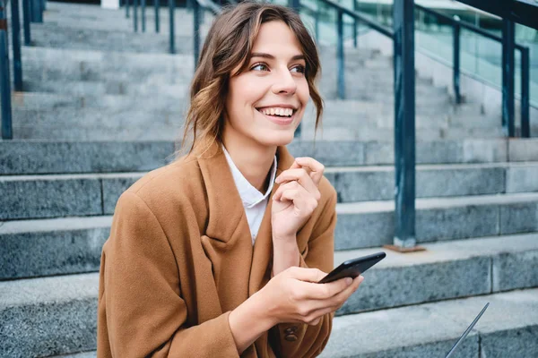 Portrett av unge smilende forretningskvinne i frakk, lykkelig ved bruk av mobiltelefon utendørs – stockfoto