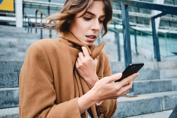 Portrait of young serious business woman fastening up coat from cold using cellphone outdoor — Stok Foto