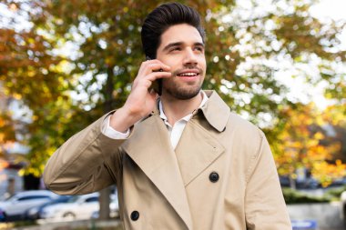 Trençkotlu genç yakışıklı adam cep telefonuyla konuşuyor, mutlu bir şekilde dışarı bakıyor.