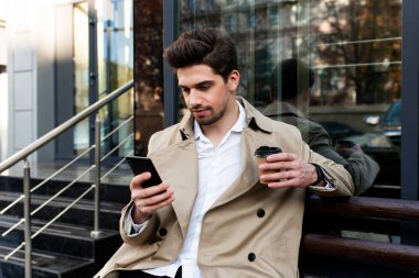 Young handsome casual man in trench coat with coffee to go thoughtfully using cellphone on city street