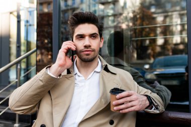 Trençkotlu ve kahveli çekici genç adam şehir caddesinde cep telefonuyla konuşuyor.