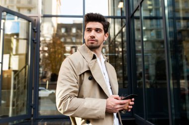 Trençkotlu ve cep telefonlu genç, yakışıklı bir adam. Düşünceli bir şekilde şehir caddesine bakıyor.