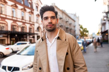 Trençkotlu çekici genç adam rüya gibi bir şekilde şehir caddesinde tek başına yürüyor.