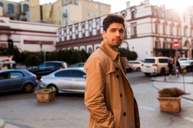 Trençkotlu çekici genç adam dikkatle kameraya bakıyor şehir caddesinde yürüyor.