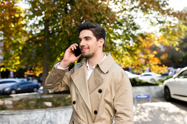 Junger attraktiver, lässiger Mann im Trenchcoat, der im Freien fröhlich auf dem Handy spricht — Stockfoto