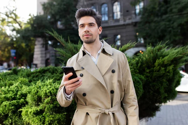 Stilvoller junger Mann im Trenchcoat mit Handy, der verträumt auf der Straße der Stadt wegschaut — Stockfoto