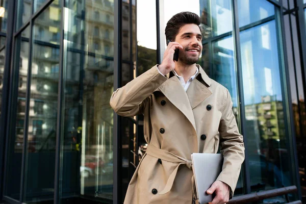 Junger gutaussehender, lässiger Geschäftsmann im Trenchcoat mit Laptop, der fröhlich auf der Straße in der Stadt mit dem Handy spricht — Stockfoto
