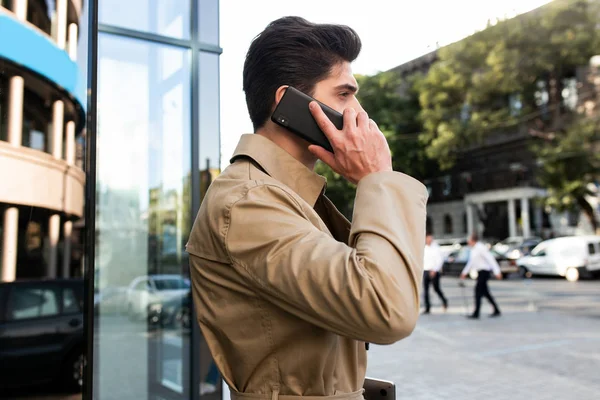 Seitenansicht eines jungen, stylischen Mannes im Trenchcoat, der auf der Straße in der Stadt mit dem Handy spricht — Stockfoto