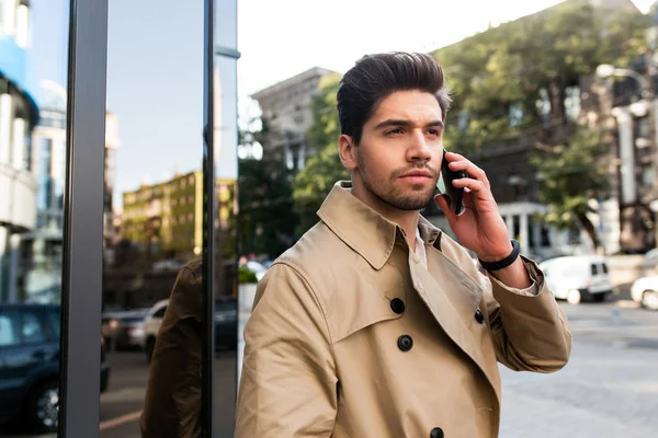 Junger stylischer Geschäftsmann im Trenchcoat, der auf der Straße der Stadt nachdenklich wegschaut und am Handy redet — Stockfoto