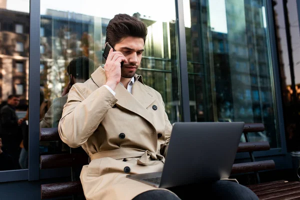 Junger lässiger Geschäftsmann im Trenchcoat arbeitet selbstbewusst am Laptop und telefoniert auf der Straße — Stockfoto