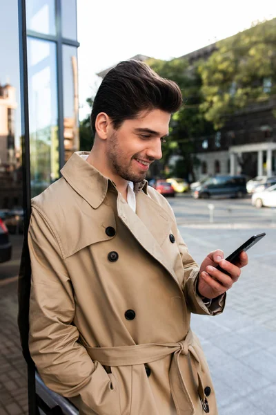 Seitenansicht eines jungen lächelnden Mannes im Trenchcoat, der auf der Straße glücklich sein Handy benutzt — Stockfoto