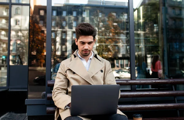 Junger attraktiver, stylischer Mann im Trenchcoat sitzt auf Bank und arbeitet nachdenklich am Laptop in der City Street — Stockfoto