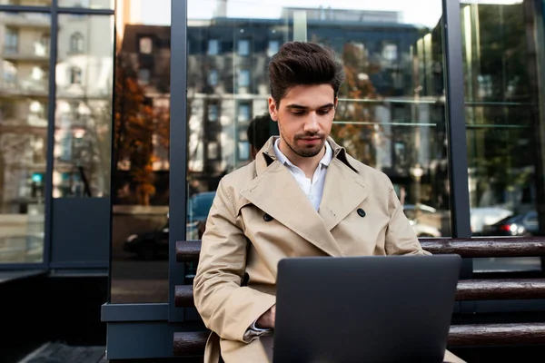 Junger hübscher stilvoller Geschäftsmann im Trenchcoat sitzt auf einer Bank und arbeitet an einem Laptop auf der Straße der Stadt — Stockfoto