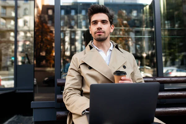 Junger attraktiver, stylischer Mann im Trenchcoat, der mit Laptop und Kaffee auf einer Bank sitzt, um nachdenklich auf der Straße der Stadt wegzuschauen — Stockfoto