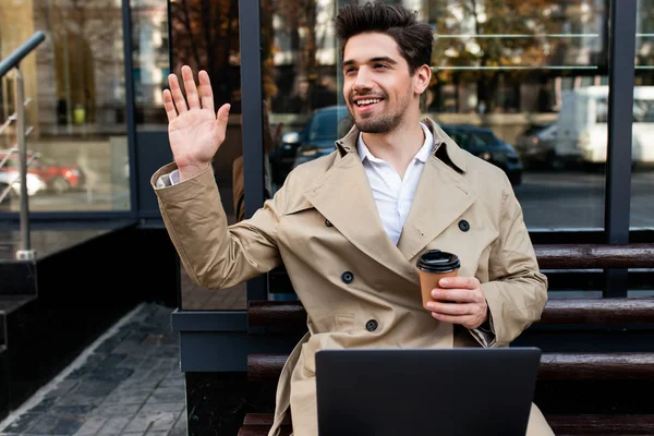 Young attractive smiling man in trench coat with laptop and coffee joyfully waving hand hello on city street