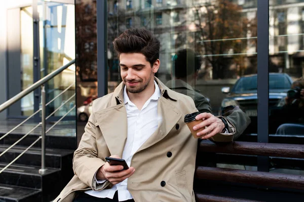 Junger lächelnder, stilvoller Mann im Trenchcoat, der auf einer Bank sitzt und Kaffee trinkt, um glücklich mit dem Handy auf der Stadtstraße zu gehen — Stockfoto