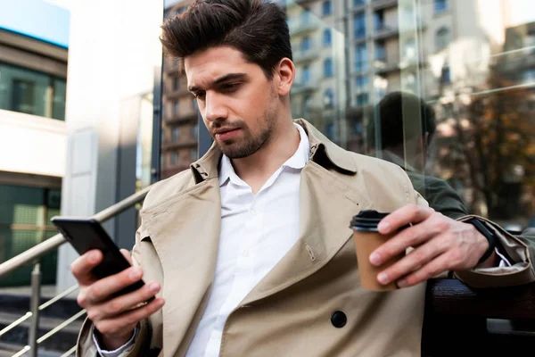 Junger selbstbewusster, lässiger Mann im Trenchcoat mit Coffee to go mit Handy auf der Stadtstraße — Stockfoto