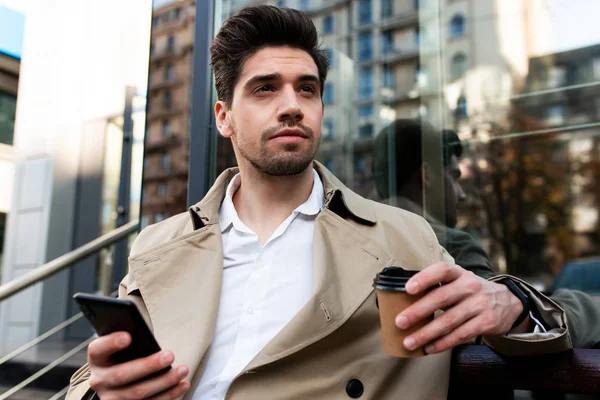 Junger lässiger Mann im Trenchcoat mit Coffee to go und Handy, der nachdenklich auf der Straße der Stadt wegschaut — Stockfoto