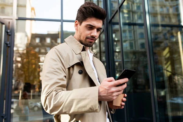 Junger attraktiver stylischer Mann im Trenchcoat mit Kaffee zum Handy-Gehen durch die Stadtstraße — Stockfoto