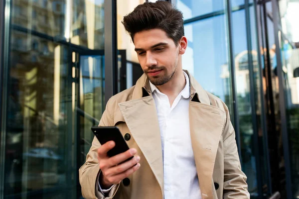 Stilvoller junger Mann im Trenchcoat mit Handy auf der Straße — Stockfoto