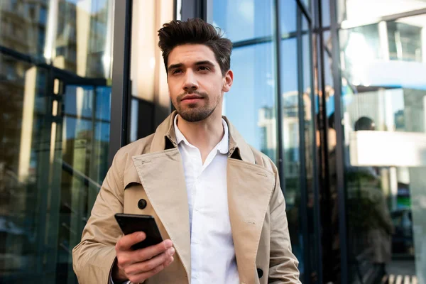 Junger attraktiver, stylischer Mann im Trenchcoat mit Handy, der auf der Straße der Stadt aufmerksam wegschaut — Stockfoto