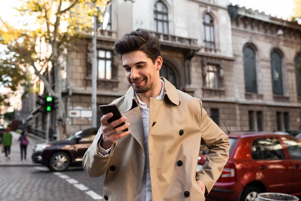 Junger attraktiver lächelnder Mann im Trenchcoat läuft glücklich mit Handy durch die Stadtstraße — Stockfoto