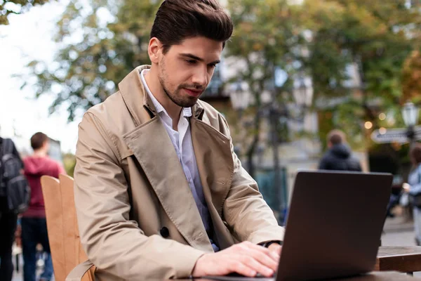 Stilvoller Geschäftsmann im Trenchcoat arbeitet selbstbewusst am Laptop in Straßencafé — Stockfoto