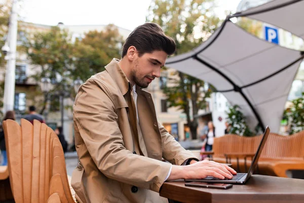 Junger gutaussehender, lässiger Geschäftsmann im Trenchcoat, der nachdenklich am Laptop in einem Café auf der Straße arbeitet — Stockfoto