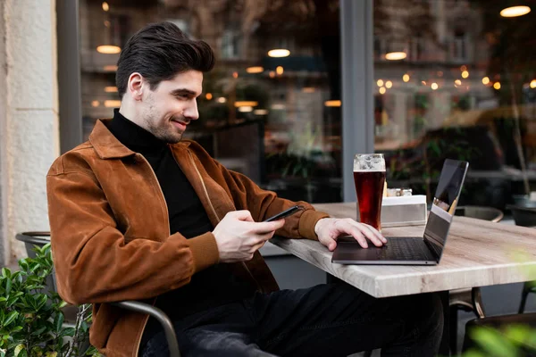 Junge attraktive Gelegenheitsunternehmer mit Handy glücklich auf Laptop arbeiten mit Bier in Café auf der Straße — Stockfoto