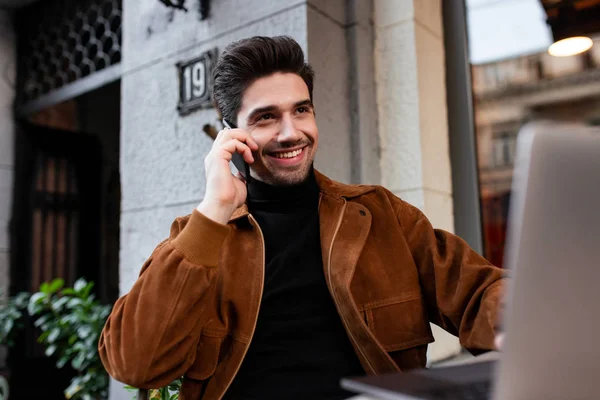 Junger fröhlicher, lässiger Mann, der fröhlich mit dem Handy telefoniert und auf dem Laptop im Café auf der Straße arbeitet — Stockfoto