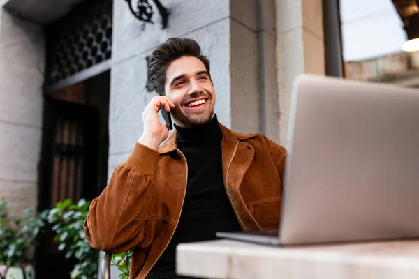 Joven sonriente hombre casual alegremente hablando en el teléfono celular trabajando en el ordenador portátil en la cafetería en la calle — Foto de Stock