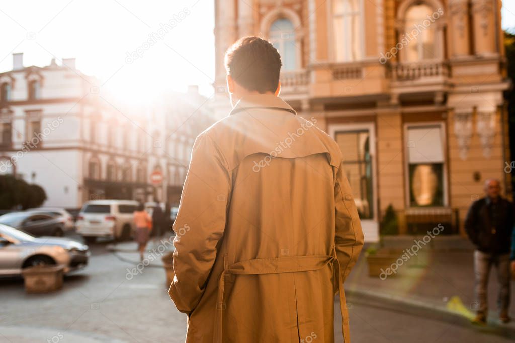 Back view of young confident man in trench coat walking through city street alone