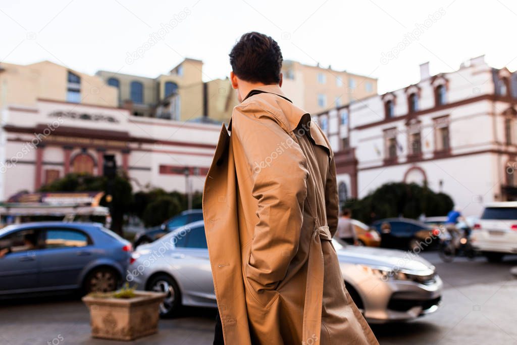 Back view of young stylish man in trench coat walking through city street 