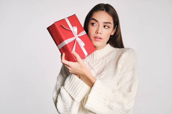 Attractive girl in cozy sweater holding gift box thoughtfully looking away over white background