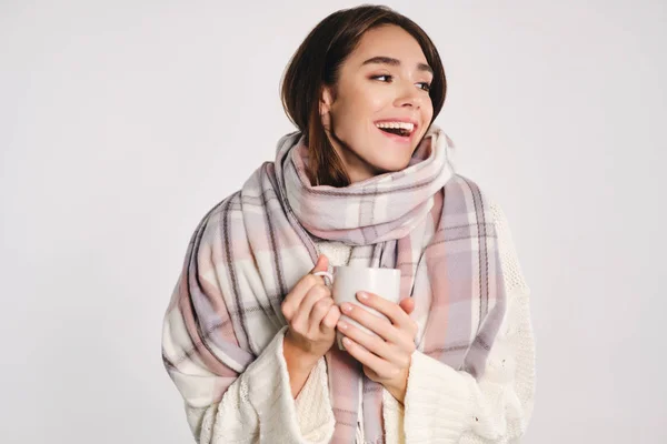 Привлекательная веселая девушка с уютным шарфом держа чашку с теплым напитком счастливо глядя в сторону на белом фоне — стоковое фото