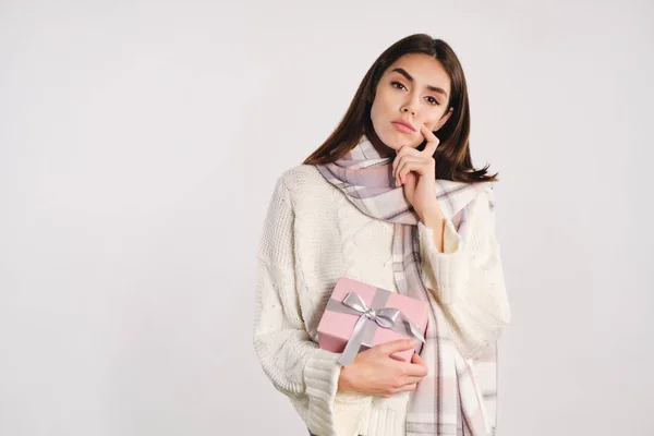 Chica pensativa en suéter acogedor con bufanda que sostiene la caja de regalo cuidadosamente mirando en cámara aislada —  Fotos de Stock