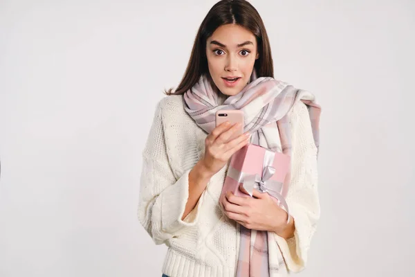 Attractive girl in cozy sweater with scarf holding gift box and cellphone surprisingly looking in camera over white background — Stock Photo, Image